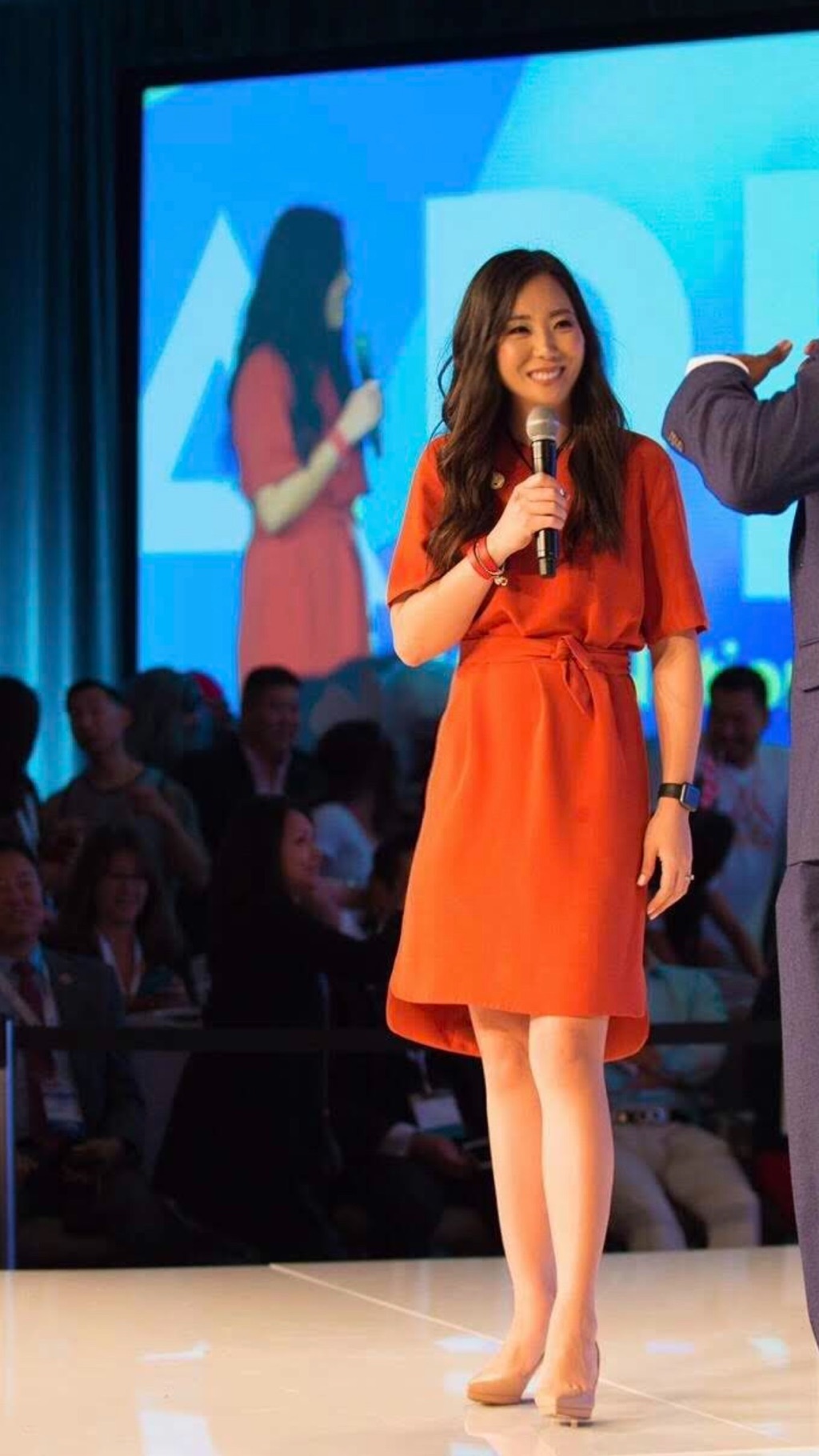Angie Lee speaking on stage in an orange dress